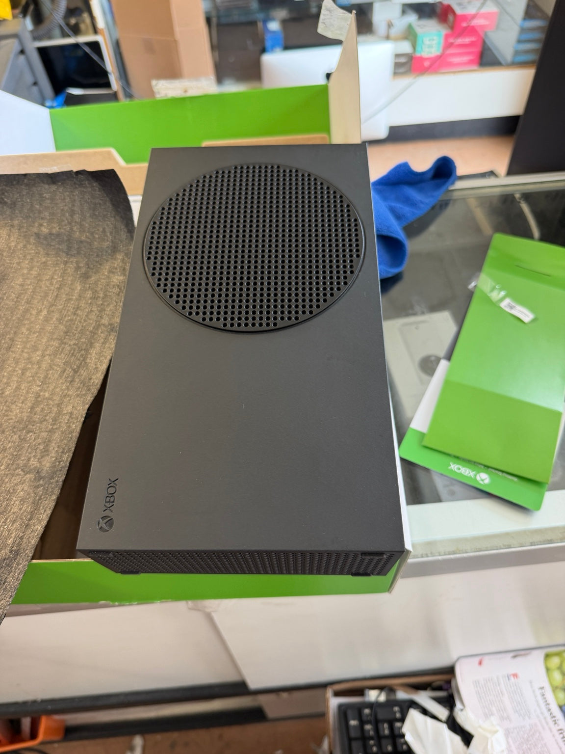 A black Xbox Series S console sitting on its green and white retail box on a glass countertop in a store setting.