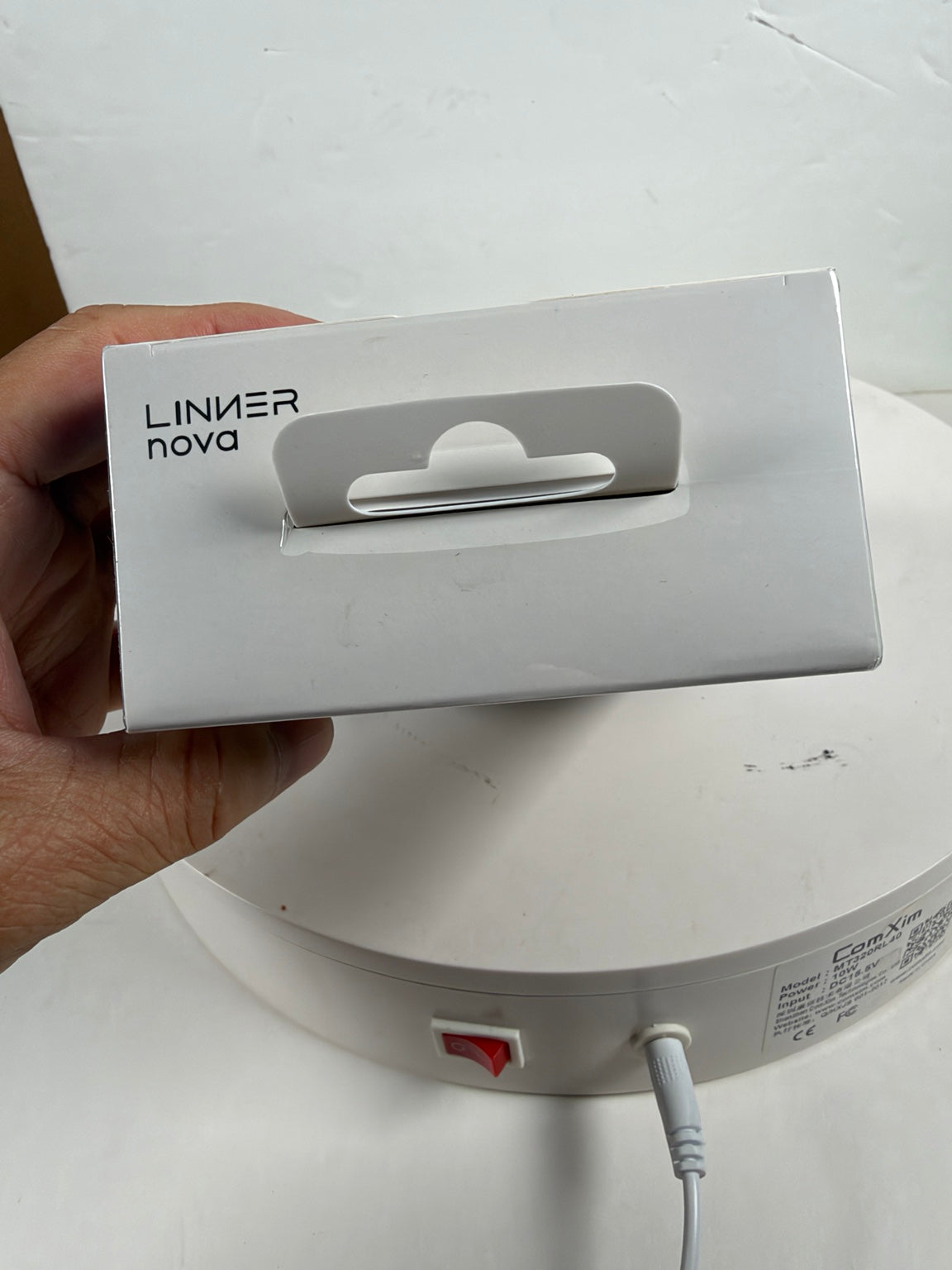 A hand holds a white Linner Nova product box in front of a white circular appliance base with a power cord plugged in.