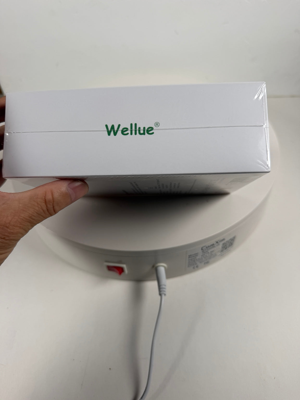 A hand holds a white Wellue product box wrapped in clear plastic, with the green 'Wellue' logo printed on the top. Below the box, a round white device with a power cord is visible against a plain light background.