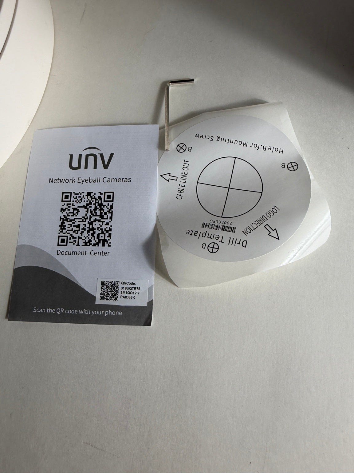 UNV network eyeball camera mounting hardware and documentation card on a light gray surface. The image shows a white circular mounting template with a metal screwdriver bit, a small hex key, and a printed UNV document card with a QR code.