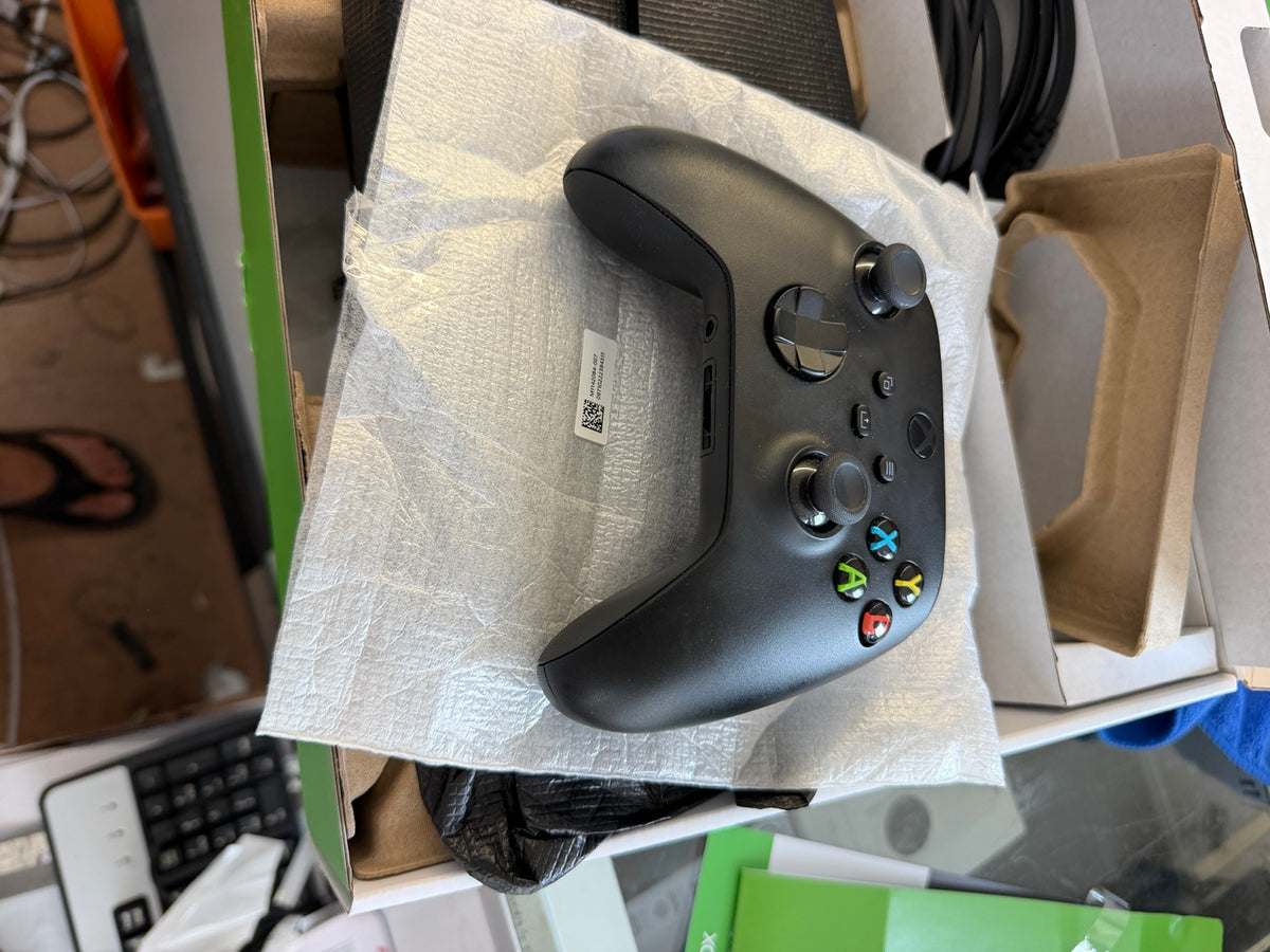 A matte black Xbox One S wireless controller resting on a crumpled piece of white paper inside an open box, surrounded by packaging materials and cables.