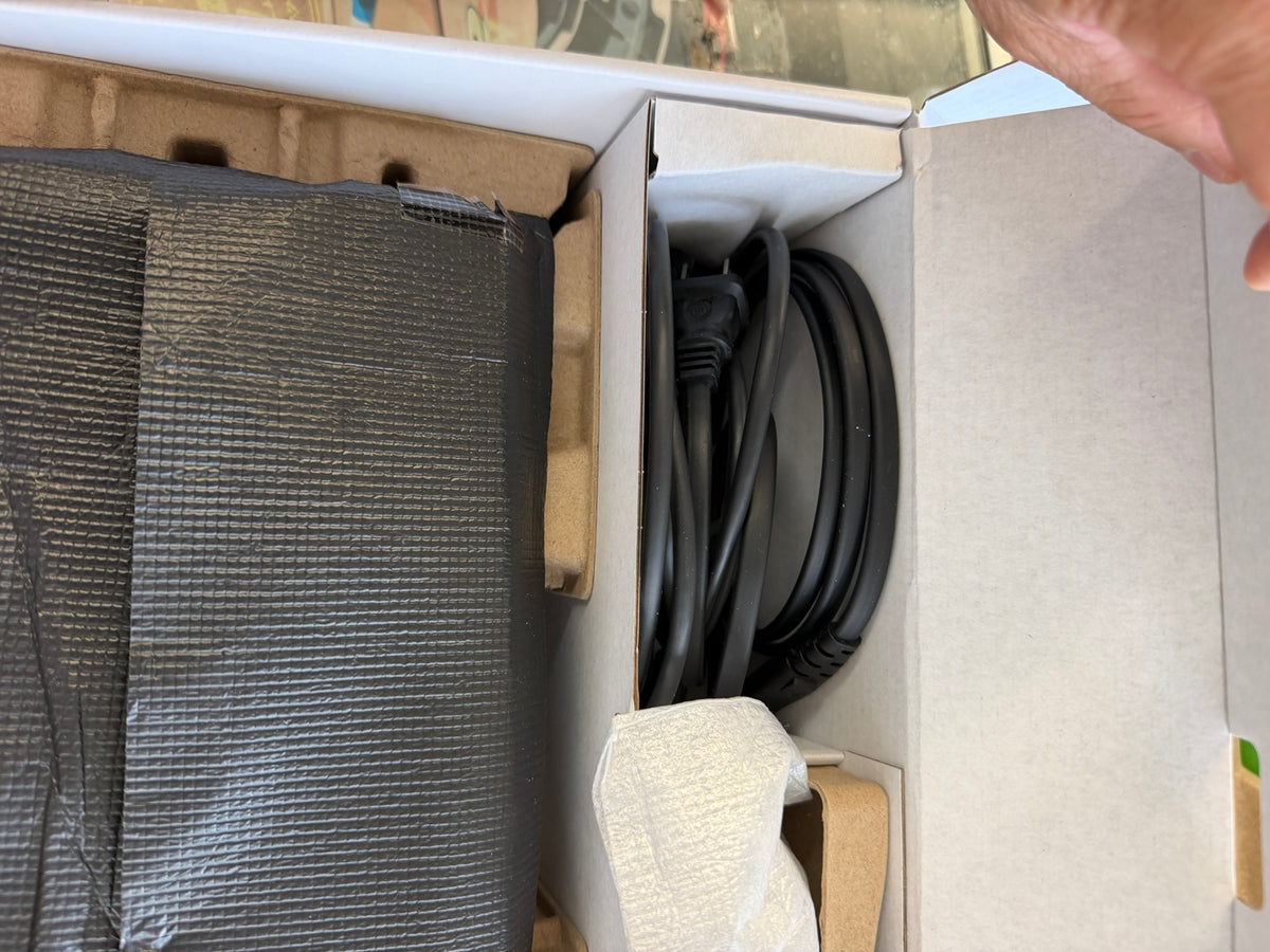 An open cardboard box showing a black textured rectangular item wrapped in protective material and a coiled black power cable inside a white box.