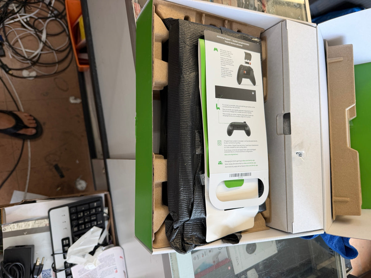 An open Xbox product box showing the contents, including a black textured accessory, a white instruction booklet with Xbox controller graphics, and cardboard inserts, photographed in a cluttered indoor setting.