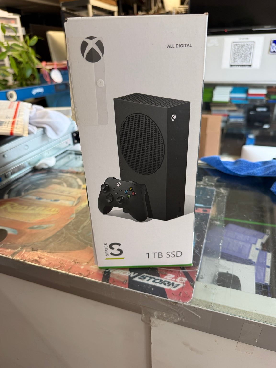 Xbox Series S 1 TB SSD game console retail box standing on a glass display counter in a store setting, with the Xbox logo and a printed image of the console and controller visible on the front.