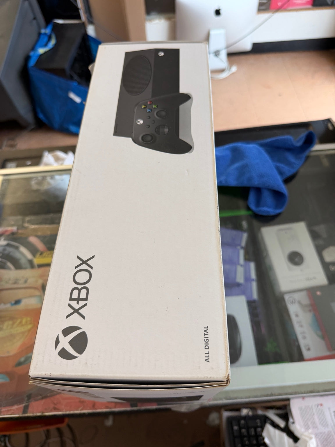 Xbox All Digital game box packaging photographed on a glass display table in a retail or office setting, with the Xbox logo and a controller graphic visible on the box.