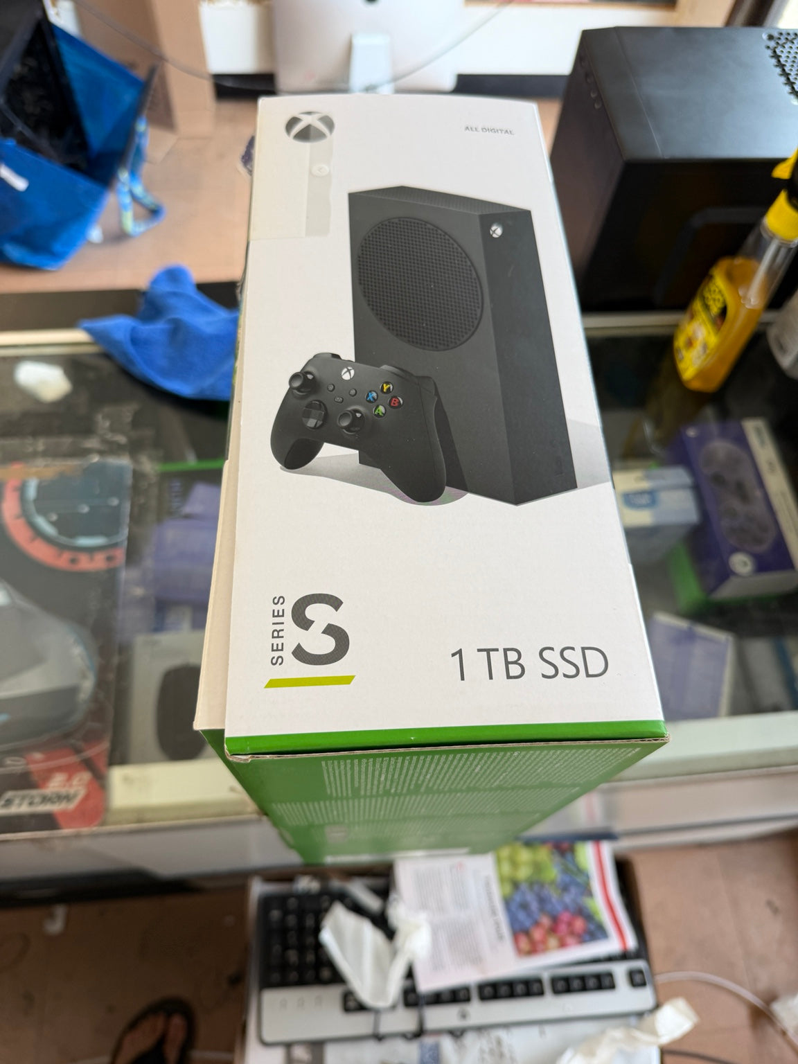 Xbox Series S 1 TB SSD retail box shown on a glass display counter in a store. The white box features a photo of the black console and controller, with green accents at the bottom.