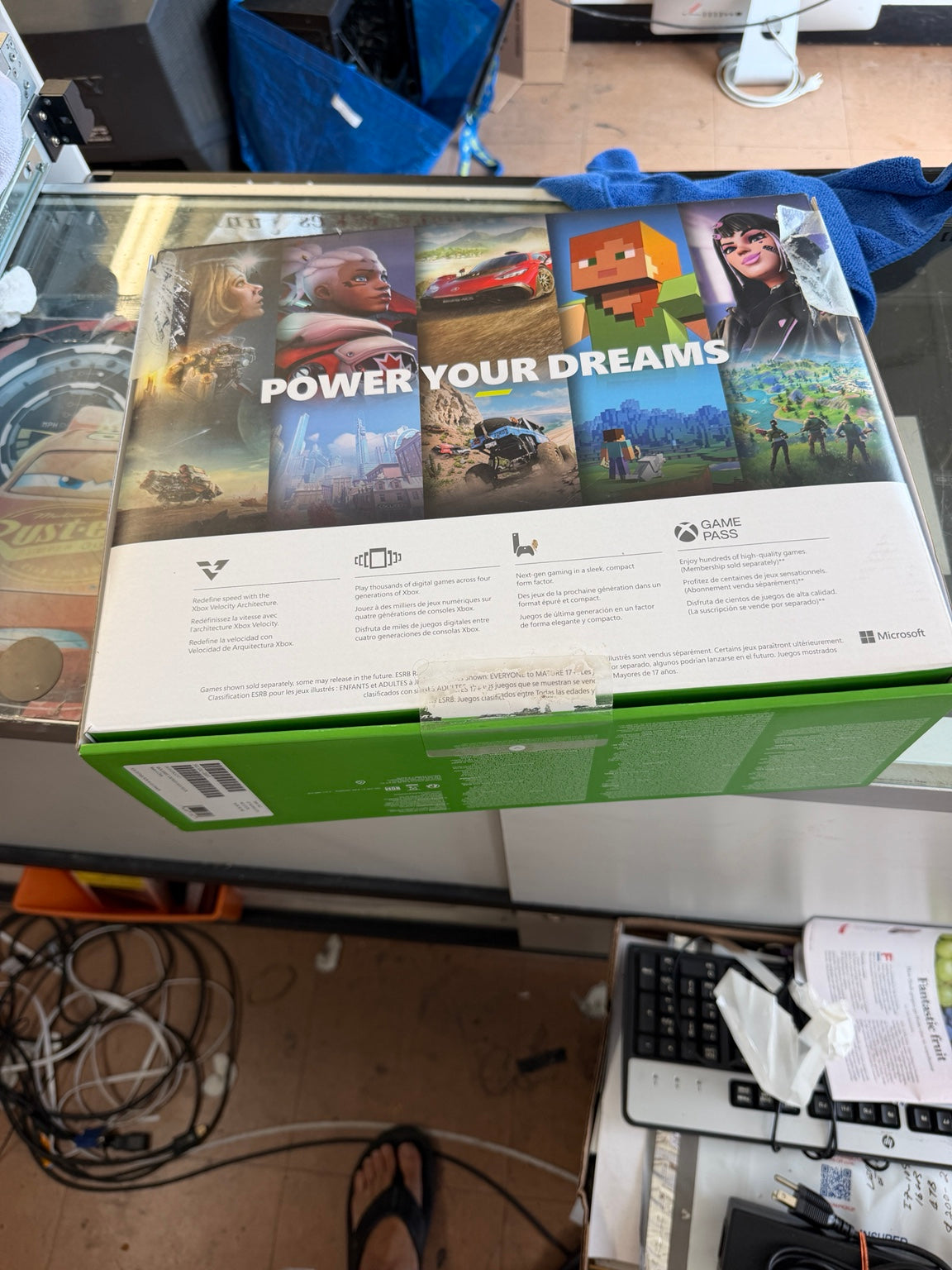 A Microsoft Xbox game box is shown from the back, resting on a glass display counter in a cluttered retail or office environment. The box features a white top section with colorful game cover art and text, and a green bottom section.