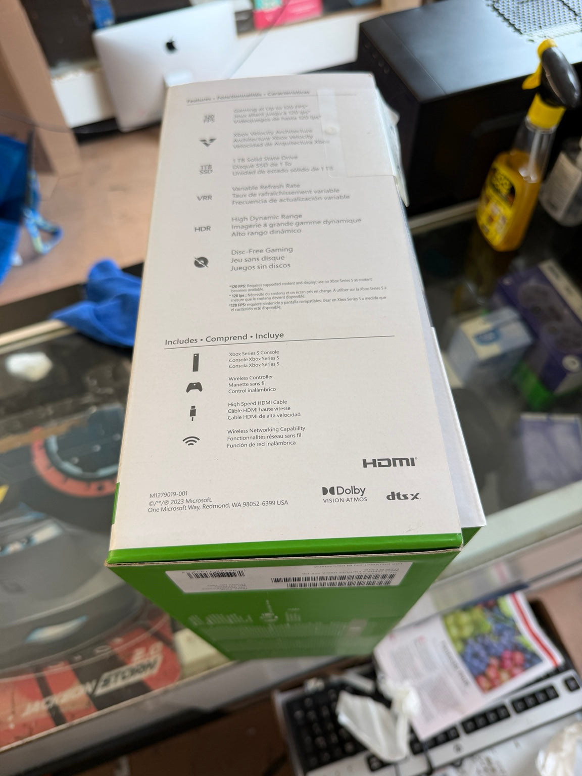A product box photographed on a glass display counter in a retail or office setting. The box is mostly white with a green band at the bottom and printed specifications, icons, and logos including HDMI, Dolby, and dts x.