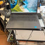 A dark gray Lenovo laptop with an open screen displaying a tropical beach wallpaper, sitting on a metal stand in a cluttered workspace with visible cables and computer parts.