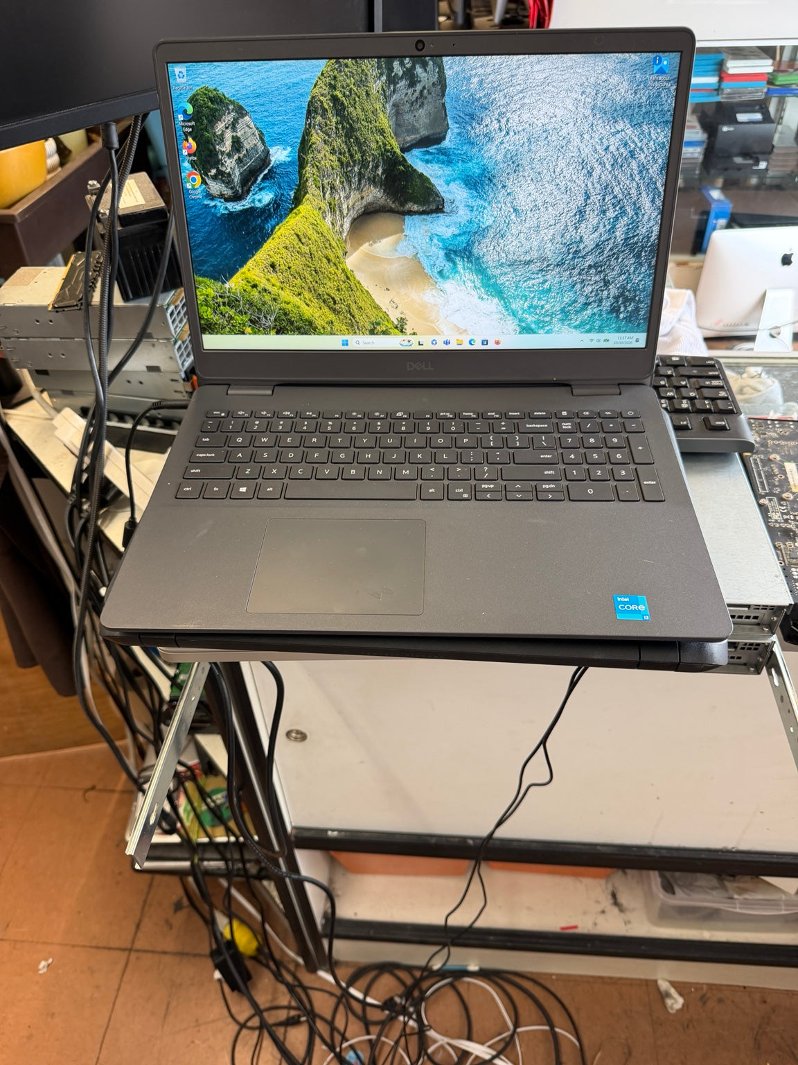 A dark gray Lenovo laptop with an open screen displaying a tropical beach wallpaper, sitting on a metal stand in a cluttered workspace with visible cables and computer parts.
