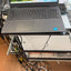 A dark gray Dell laptop with an open screen displaying a beach landscape wallpaper, resting on a metal stand in a cluttered workspace with visible cables and other equipment.