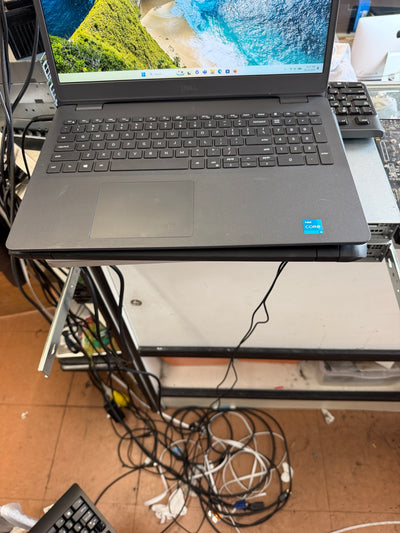 A dark gray Dell laptop with an open screen displaying a beach landscape wallpaper, resting on a metal stand in a cluttered workspace with visible cables and other equipment.