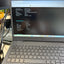 A Dell laptop open on a desk, showing the battery health information screen on the display. The laptop is gray with black keys, and the background shows a cluttered office environment with other monitors and cables.