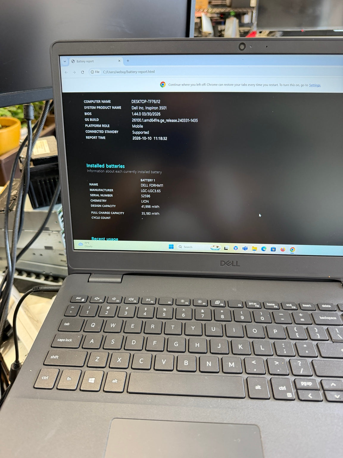 A Dell laptop open on a desk, showing the battery health information screen on the display. The laptop is gray with black keys, and the background shows a cluttered office environment with other monitors and cables.