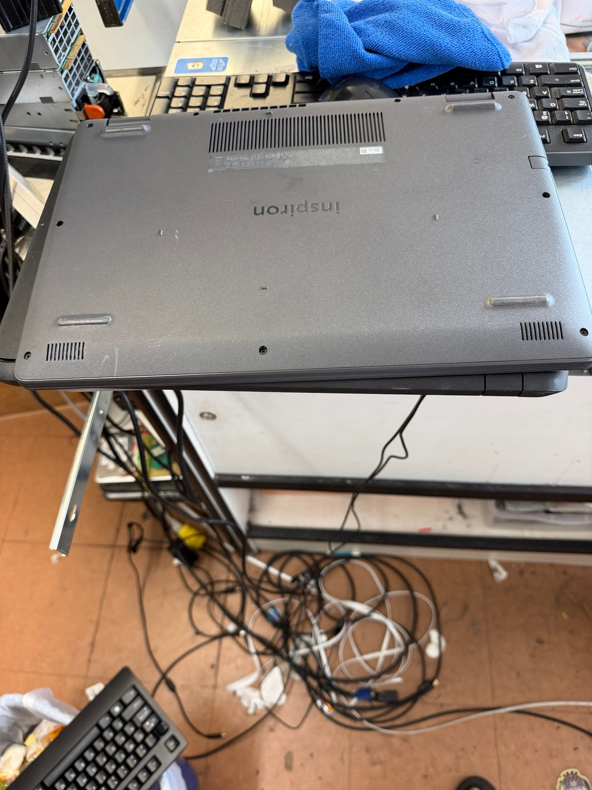 A gray Lenovo Inspiron laptop shown from the underside, resting on a stand in a cluttered workspace with keyboards, cables, and a blue cloth visible.