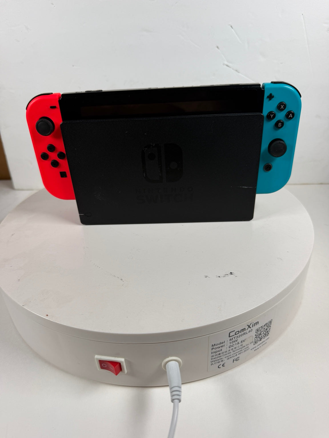 Nintendo Switch console docked in a black charging stand, with red and blue Joy-Con controllers attached, set on a white round charging pedestal against a white wall.