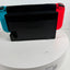 Nintendo Switch with a black dock or case in the center, featuring a turquoise left Joy-Con and a red right Joy-Con, displayed on a white round stand against a white wall.