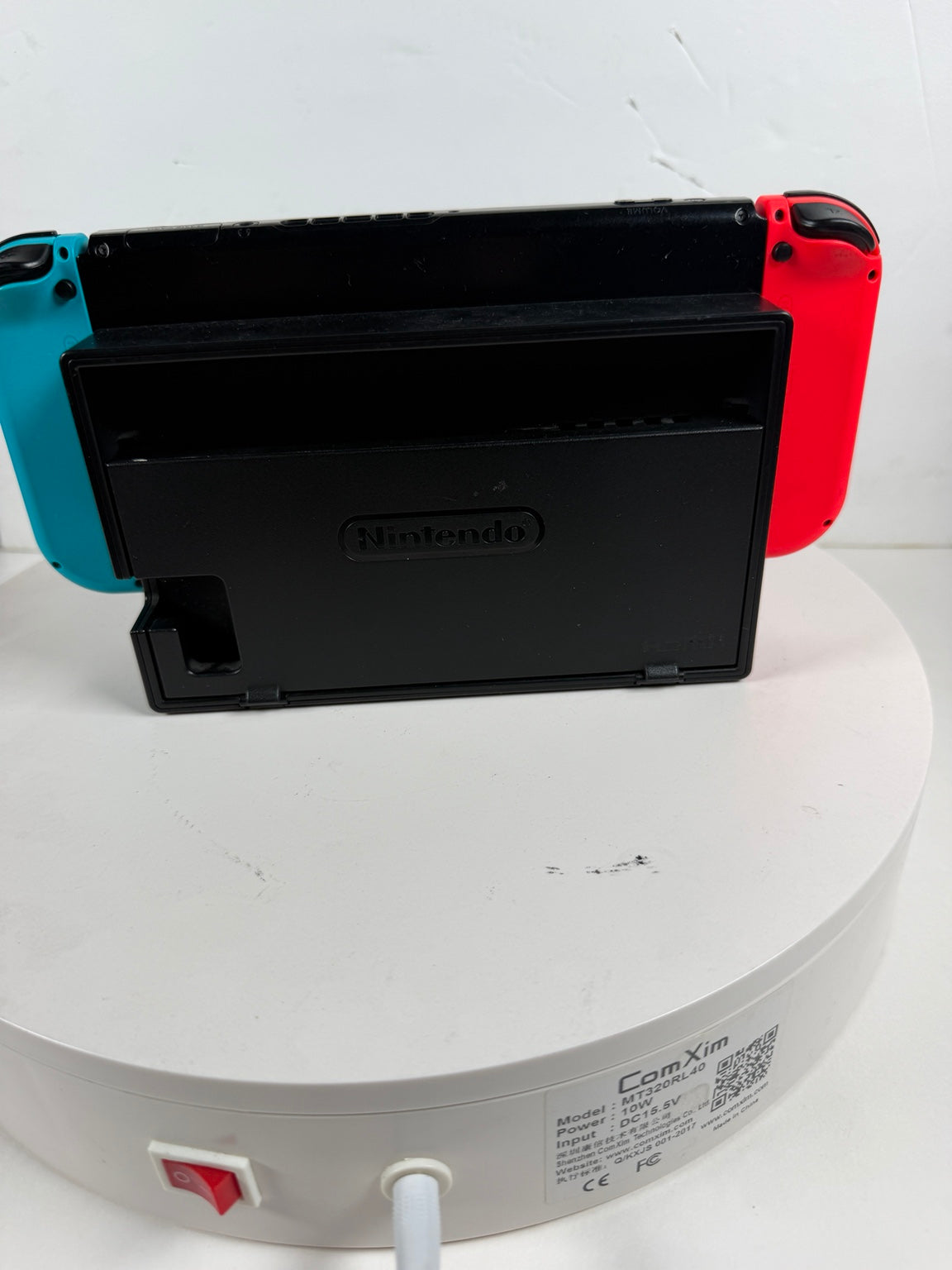 Nintendo Switch with a black dock or case in the center, featuring a turquoise left Joy-Con and a red right Joy-Con, displayed on a white round stand against a white wall.