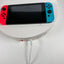 Nintendo Switch Lite handheld gaming console with a red left Joy-Con and a blue right Joy-Con attached, resting on a white round display stand with a white charging cable plugged in.