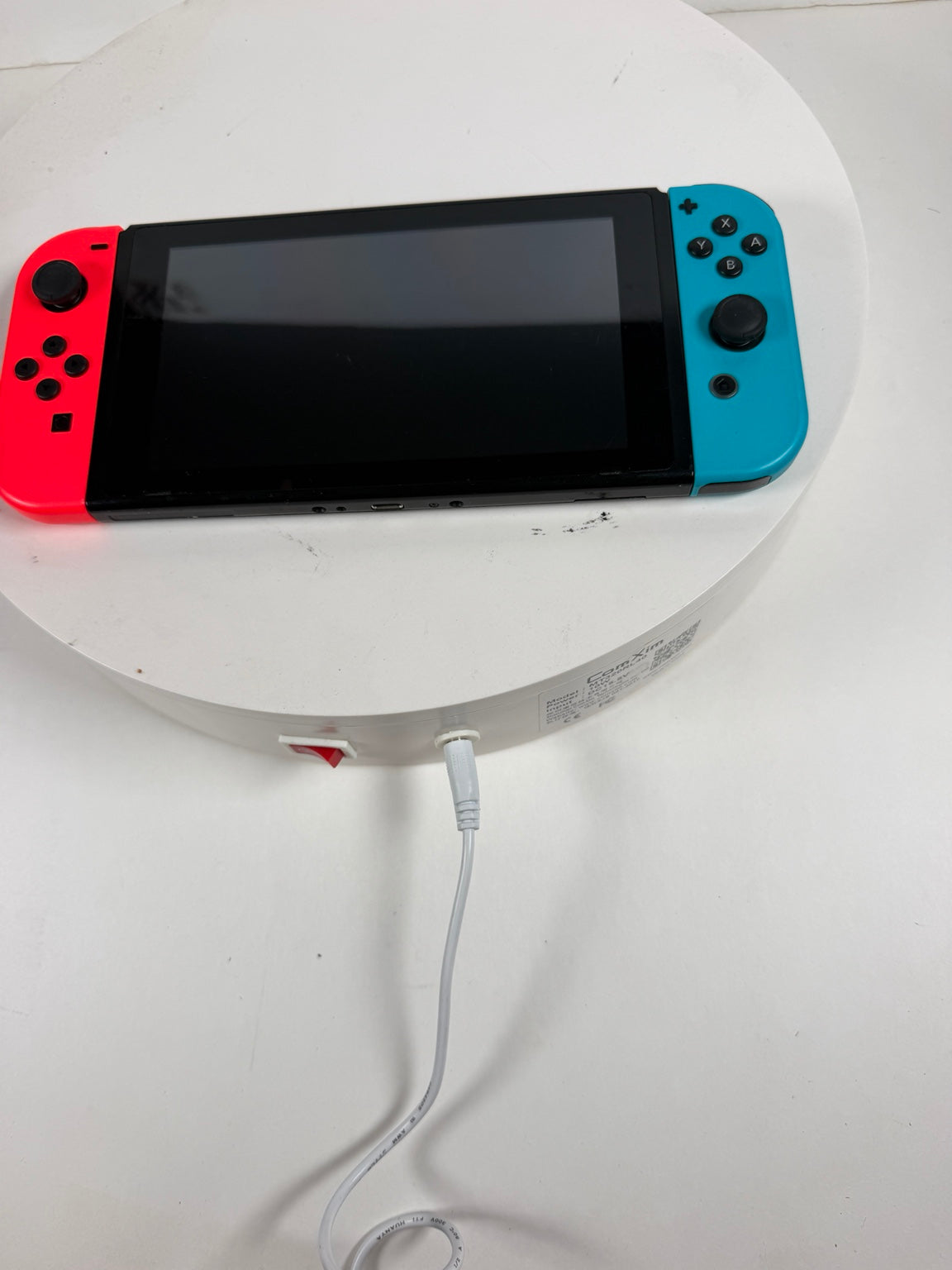Nintendo Switch Lite handheld gaming console with a red left Joy-Con and a blue right Joy-Con attached, resting on a white round display stand with a white charging cable plugged in.
