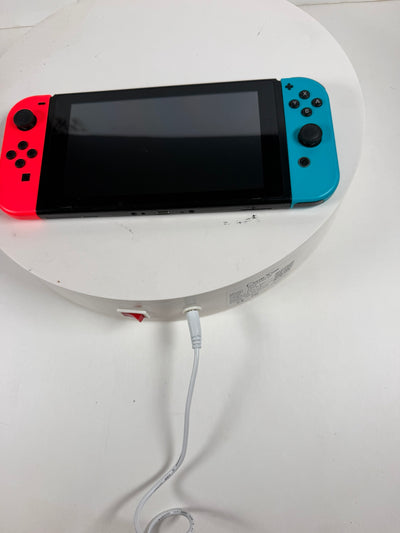 Nintendo Switch Lite handheld gaming console with a red left Joy-Con and a blue right Joy-Con attached, resting on a white round display stand with a white charging cable plugged in.