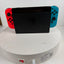 Nintendo Switch console docked in a black charging stand, with red and blue Joy-Con controllers attached, set on a white round charging pedestal against a white wall.