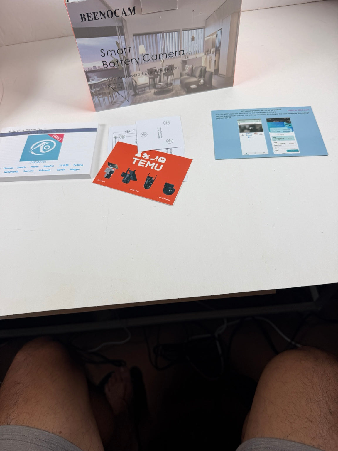 A person's legs are visible at the bottom of the frame, looking down at a white desk surface containing a Beenocam Smart Battery Camera box, a blue instruction booklet, a red TEMU card, and a white card with a blue logo.