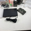 A solar-powered security camera kit laid out on a white table, including a black solar panel with cable, a camouflage-patterned camera unit, a separate camouflage bracket or accessory, packaging materials, and a plastic bag. A person's legs are visible at the bottom of the frame.