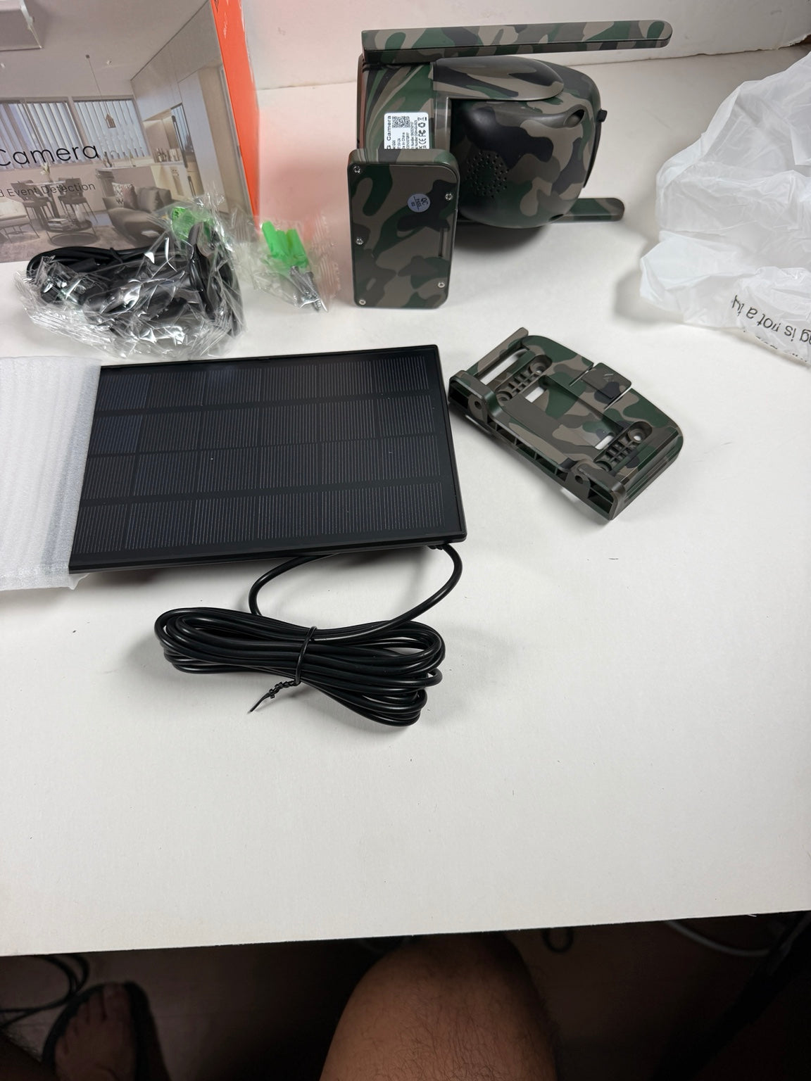 A solar-powered security camera kit laid out on a white table, including a black solar panel with cable, a camouflage-patterned camera unit, a separate camouflage bracket or accessory, packaging materials, and a plastic bag. A person's legs are visible at the bottom of the frame.