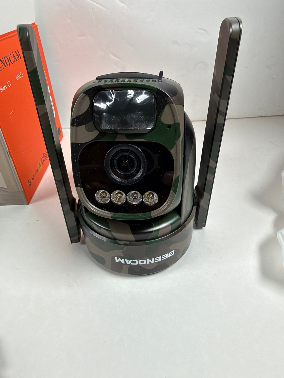 A BEENOCAM wireless security camera with a black and green camouflage pattern, featuring two vertical antennas and a clear face shield, shown on a white surface next to its orange retail box.