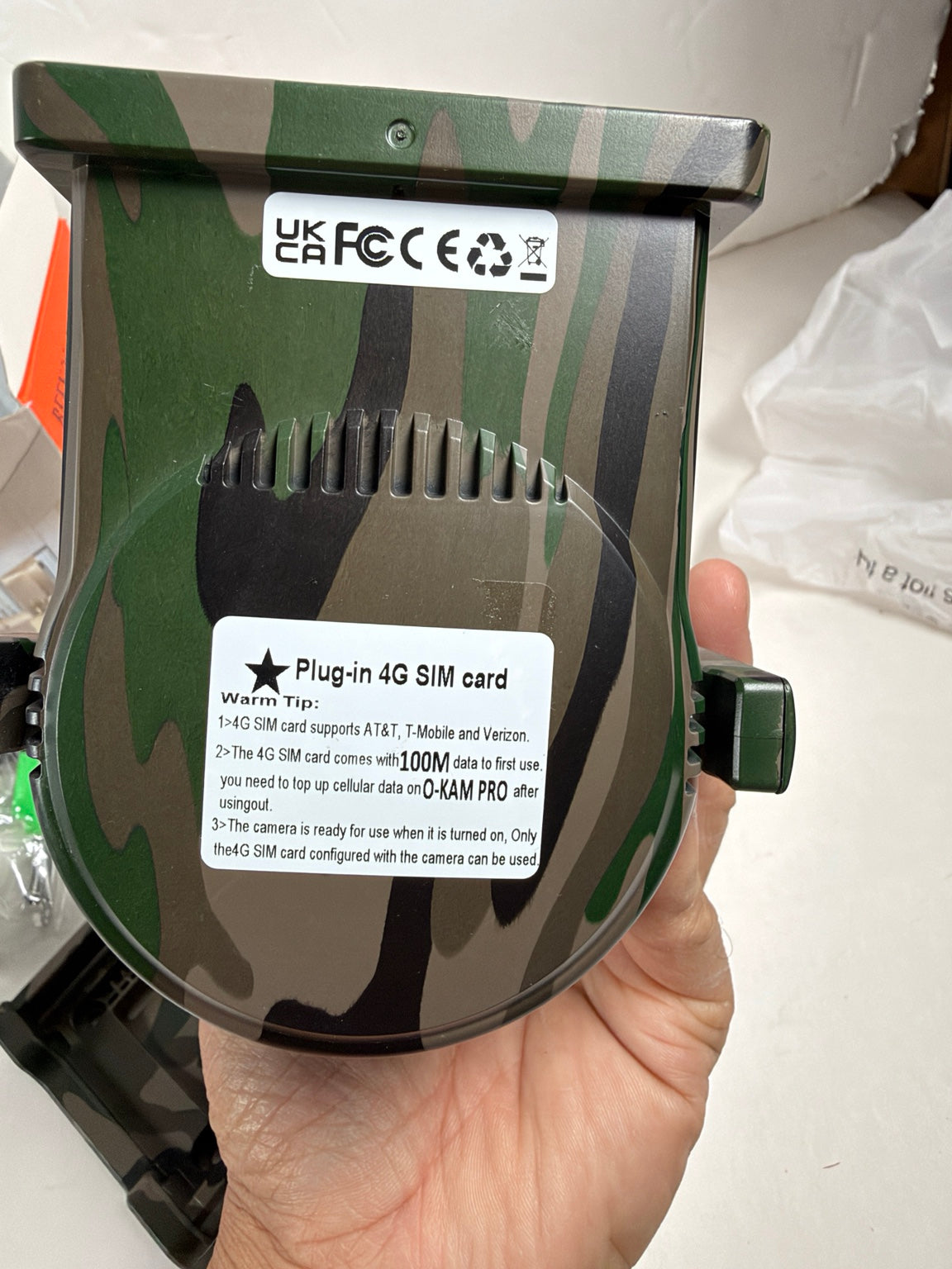 A hand holding a green camouflage-patterned UK plug-in 4G SIM card adapter device, showing the back with UK FC/CE certification stickers and a white instruction label.