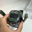 A hand holding a camouflage-patterned electronic device with two side-mounted handles, photographed against a white surface.