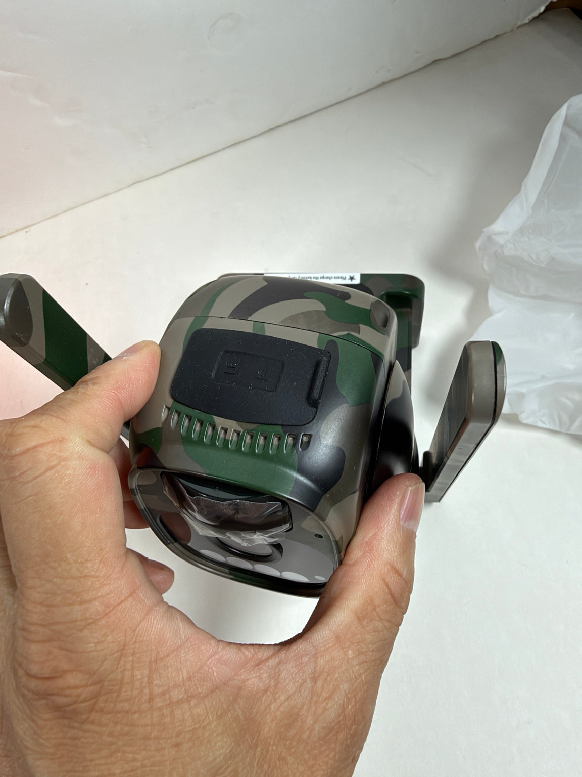 A hand holding a camouflage-patterned electronic device with two side-mounted handles, photographed against a white surface.