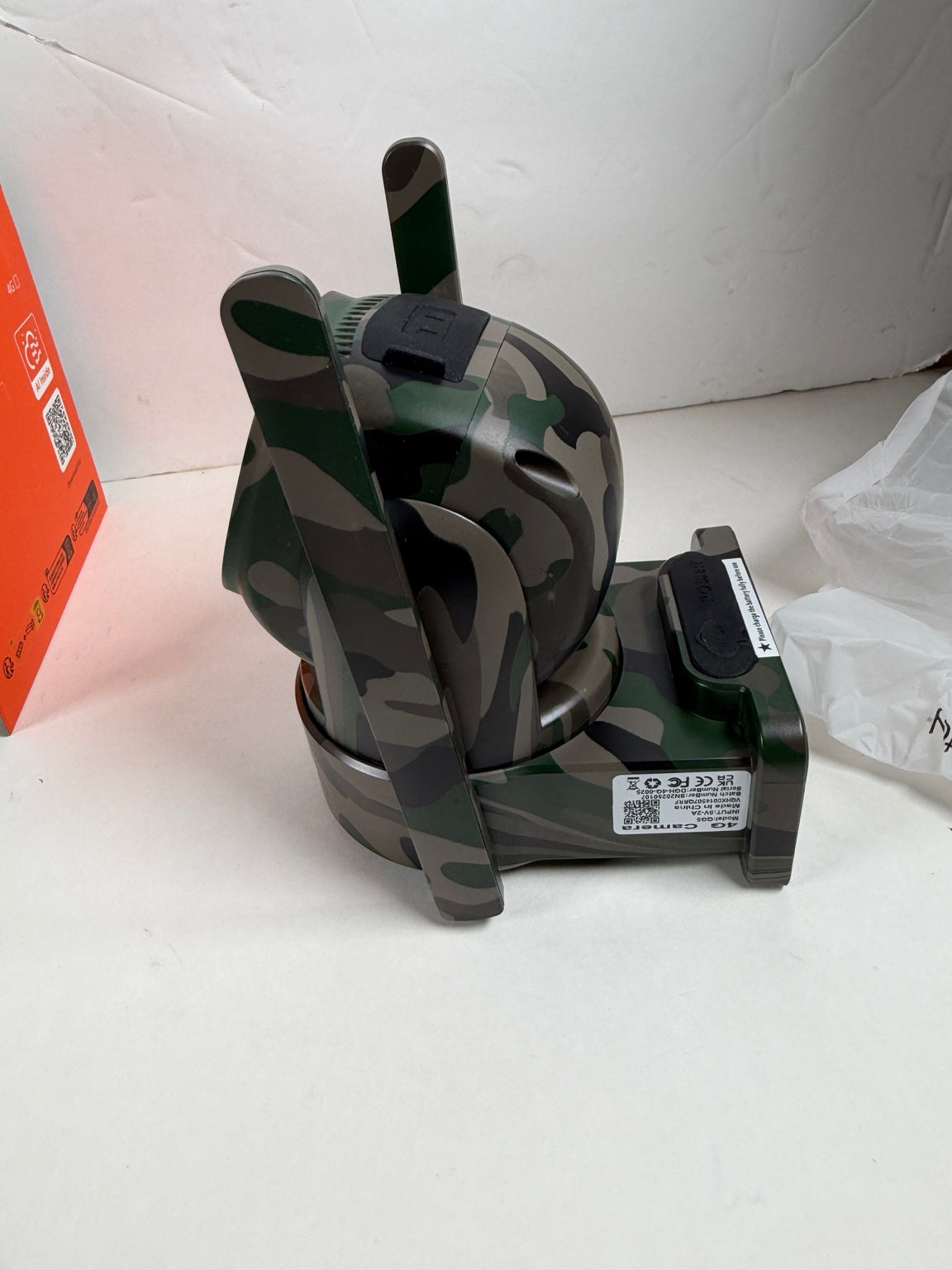 A camouflage-patterned electronic device with two antennas and a black base, shown on a white surface with its packaging partially visible.