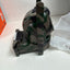 A camouflage-patterned camera lens protector or lens cover with a black mounting base, shown on a white surface next to its orange packaging box and a white plastic bag.