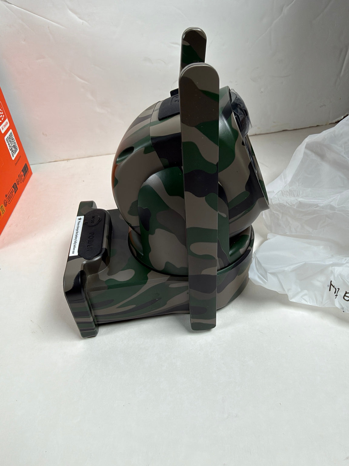 A camouflage-patterned camera lens protector or lens cover with a black mounting base, shown on a white surface next to its orange packaging box and a white plastic bag.