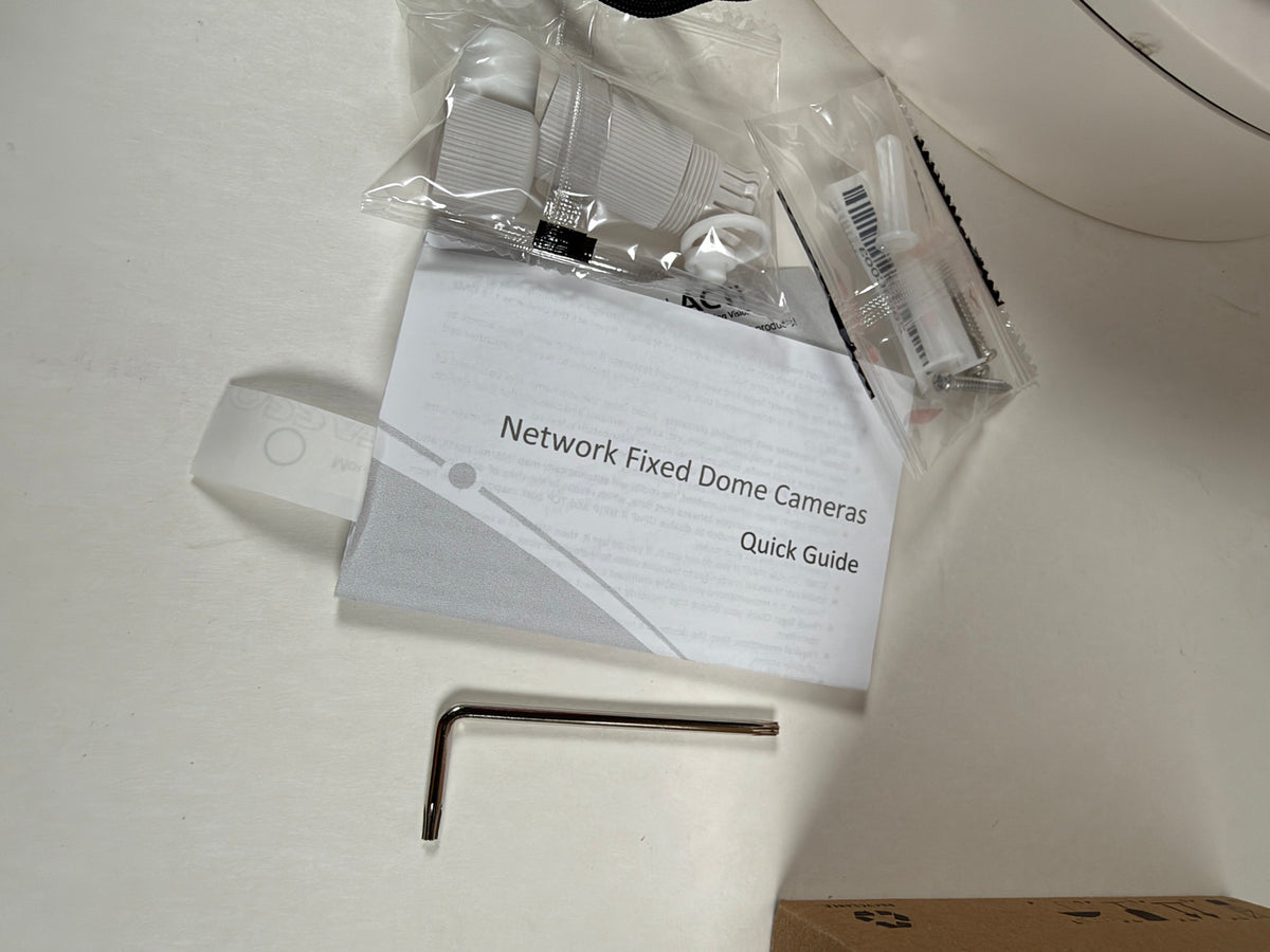Network fixed dome camera kit contents laid out on a white surface, including a quick guide booklet, mounting hardware in a clear plastic bag, and a metal hex key.