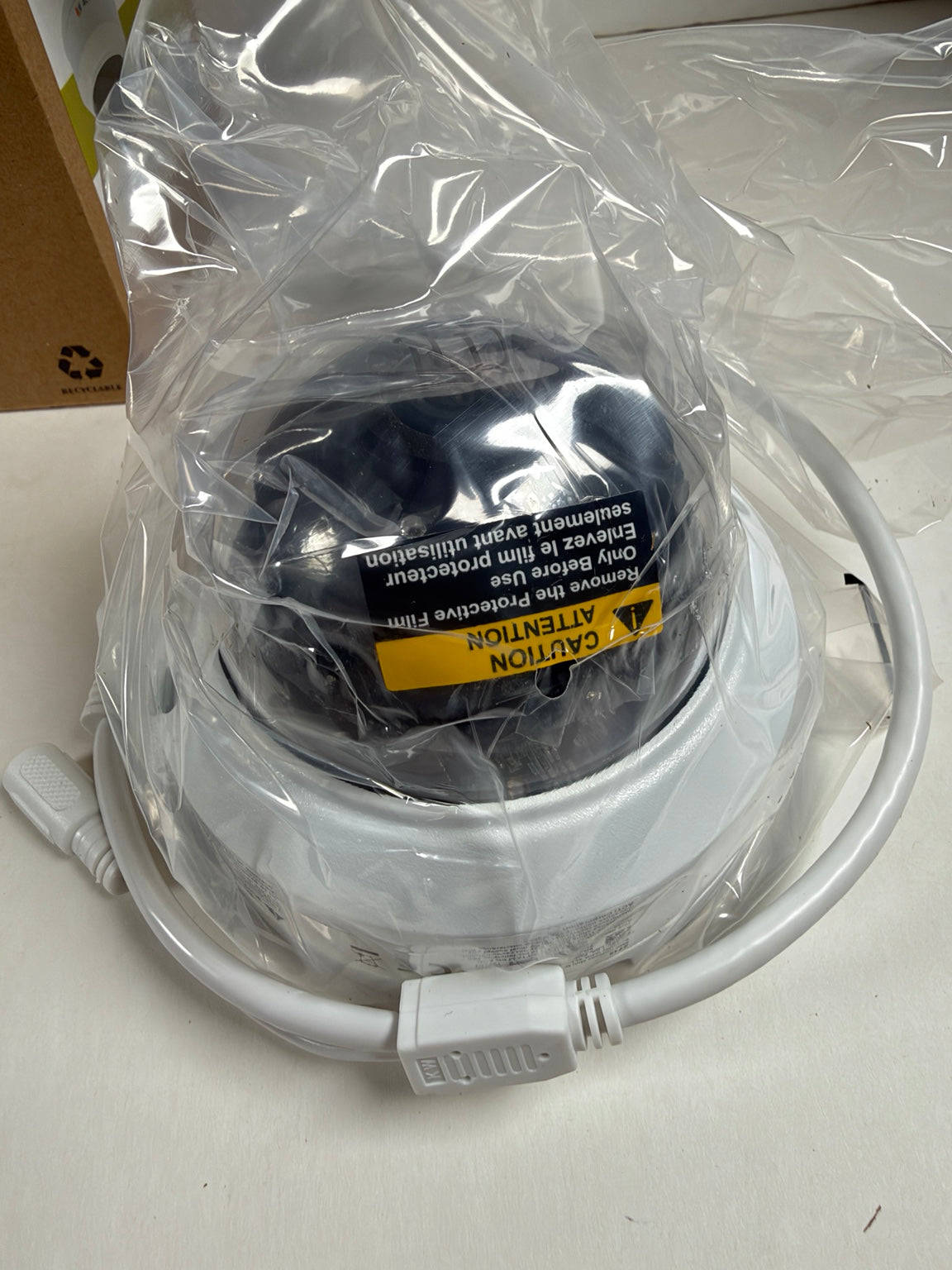 A white dome security camera still in its clear plastic packaging, showing the protective film and a yellow warning label on the front.