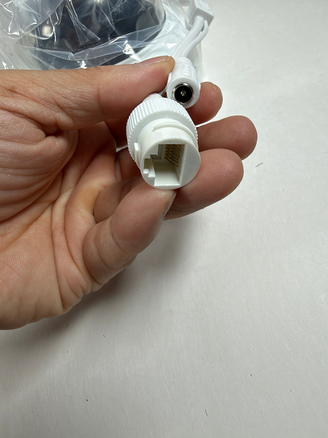 A hand holding a white plastic RJ45 connector with a short cable attached, with a plastic-wrapped item in the background on a light surface.