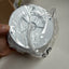 A white circular plastic component, likely a ceiling fan canopy or light fixture part, wrapped in clear plastic packaging and held by a hand against a light gray background.