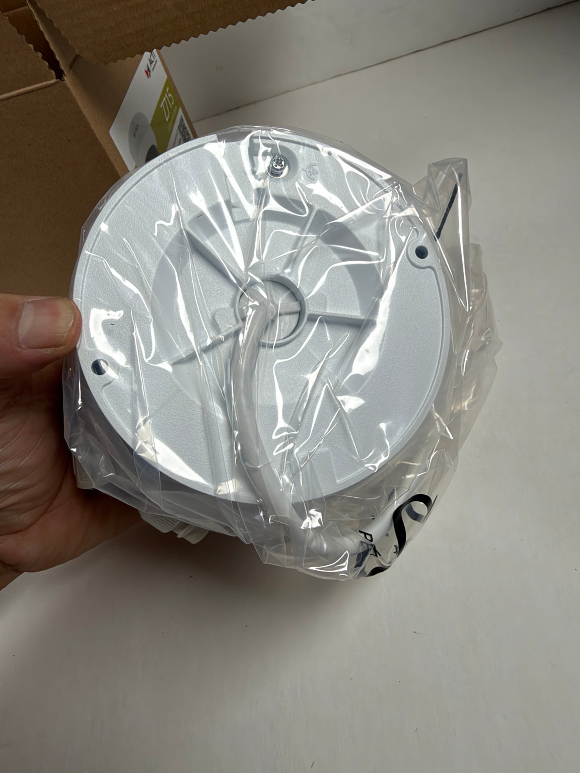 A white circular plastic component, likely a ceiling fan canopy or light fixture part, wrapped in clear plastic packaging and held by a hand against a light gray background.