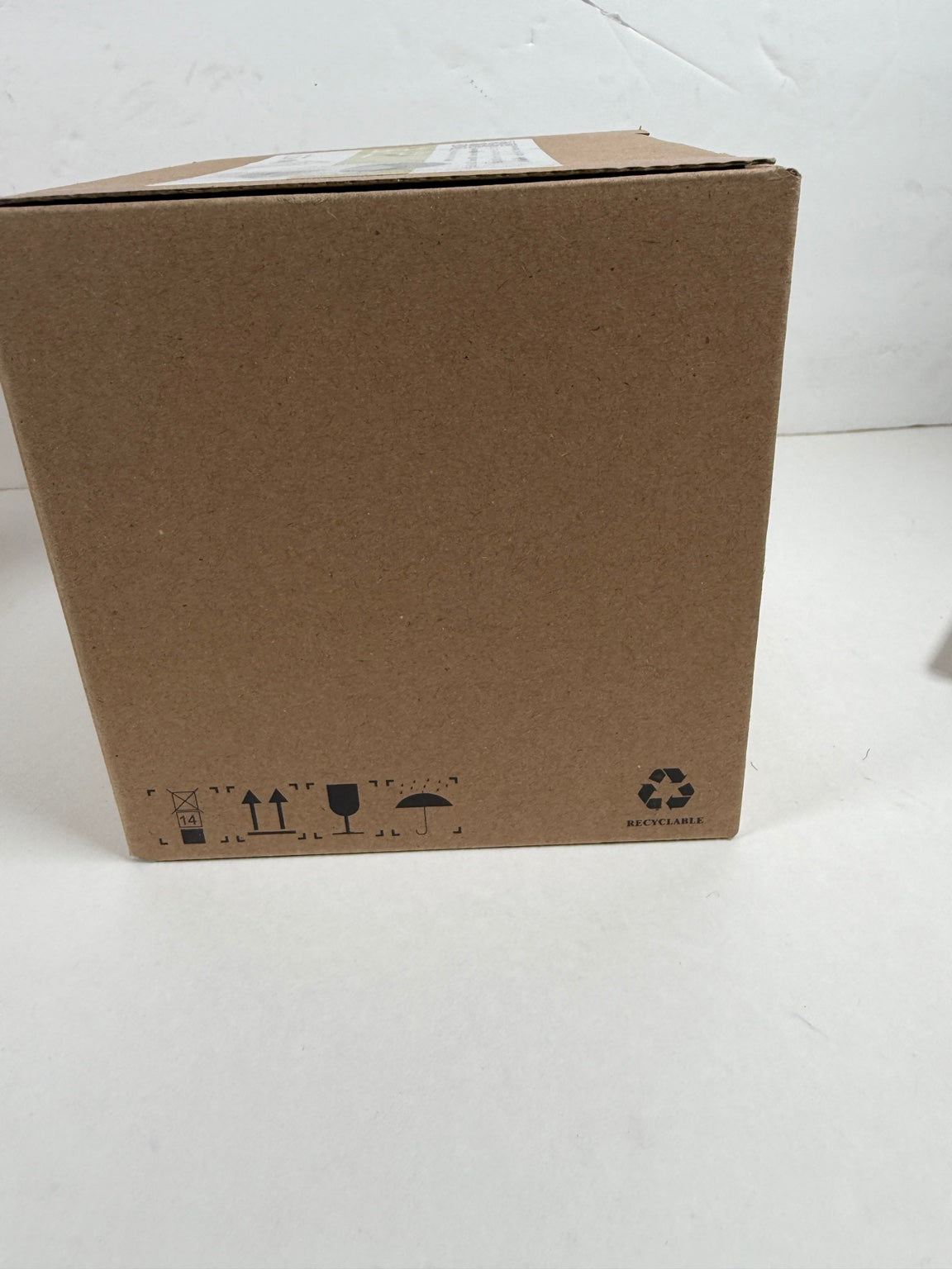 A plain brown cardboard box photographed on a white surface against a white wall, with handling icons and a recycling symbol printed near the bottom.