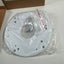 A white circular mounting plate or lens cover accessory, still sealed in clear plastic packaging with a few screws inside, placed on a light surface with a cardboard box in the background.
