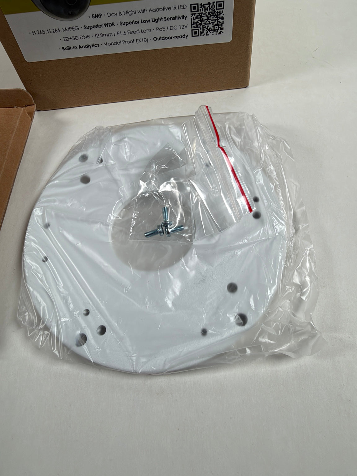 A white circular mounting plate or lens cover accessory, still sealed in clear plastic packaging with a few screws inside, placed on a light surface with a cardboard box in the background.