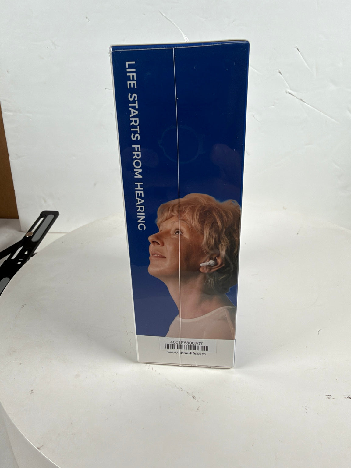 A tall rectangular product box standing upright on a white surface against a white wall. The box is predominantly dark blue with vertical white text reading 'LIFE STARTS FROM HEARING' and a printed photo of a woman wearing a small ear device.