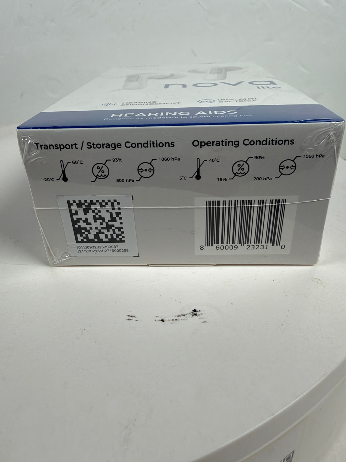 A white and blue cardboard box of Nova Hearing aids, showing the back side with text about transport and operating conditions, a QR code, and a barcode.