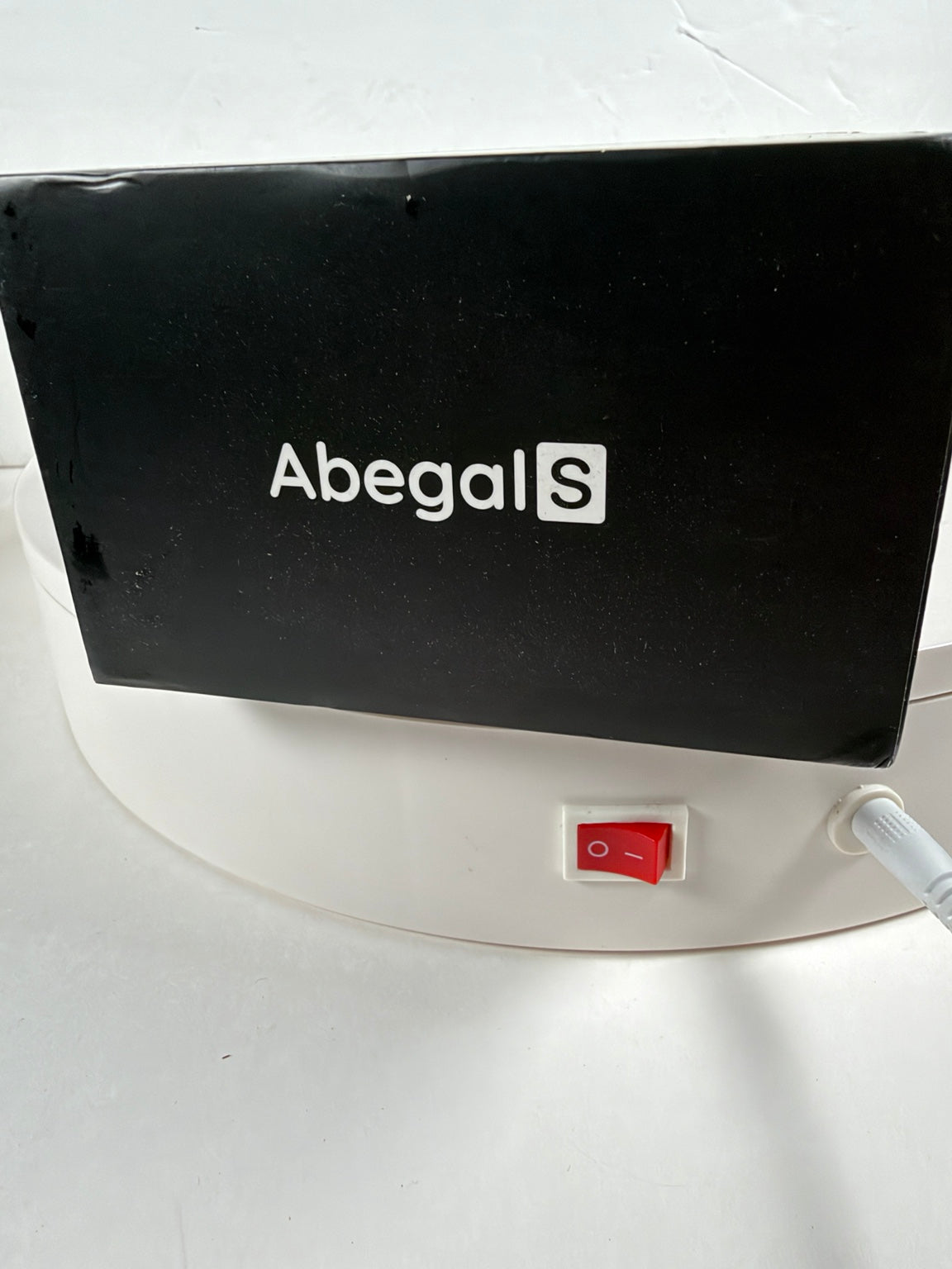 A close-up of a black rectangular device with the white AbegalS logo, mounted on a white circular base with a red power switch and a white tube connected to it.
