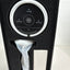 A black circular facial toning head with a silver outer ring, displayed inside its black foam presentation box. The box also contains a white-handled device wrapped in plastic and a white rectangular accessory.