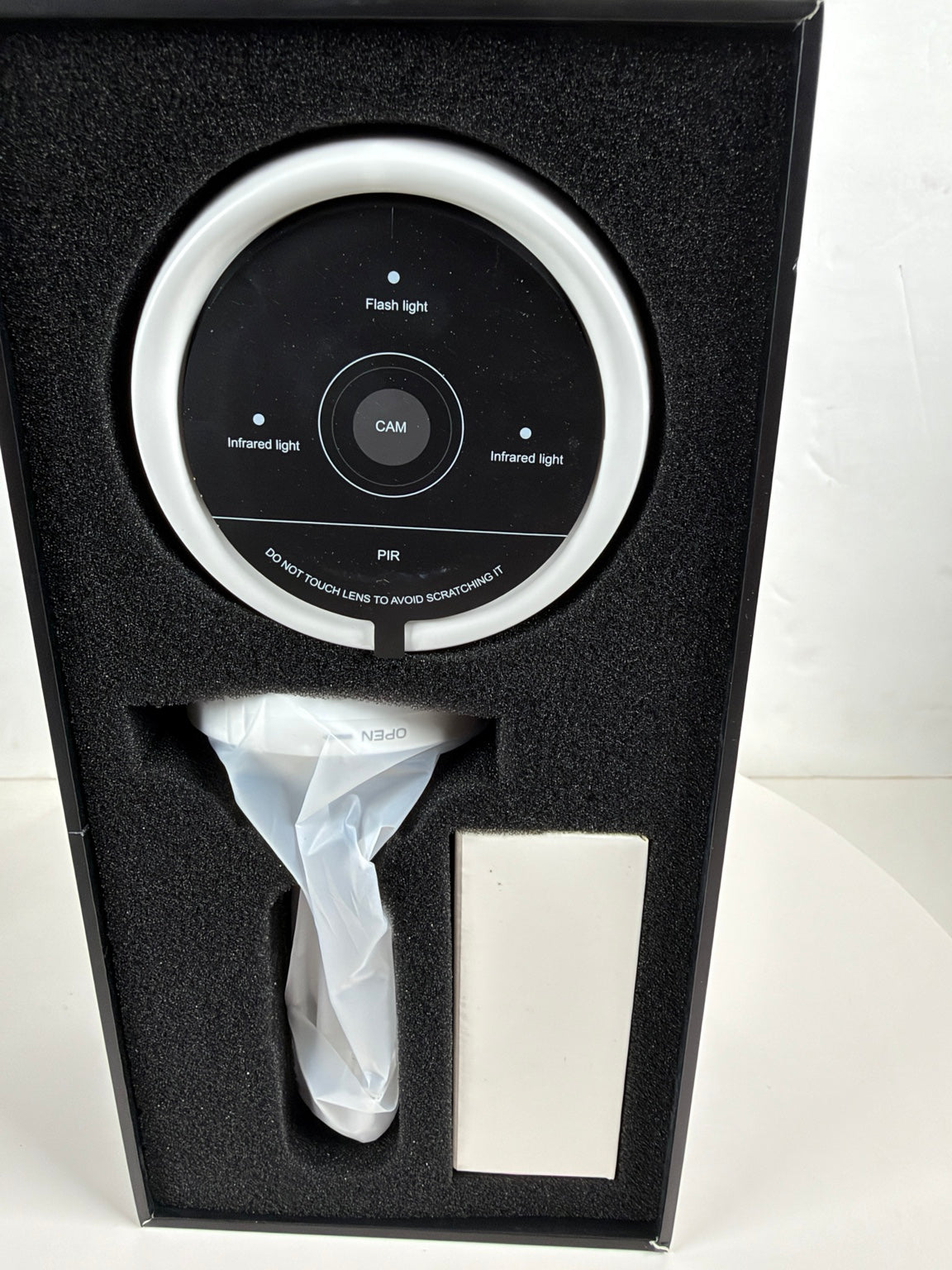 A black circular facial toning head with a silver outer ring, displayed inside its black foam presentation box. The box also contains a white-handled device wrapped in plastic and a white rectangular accessory.