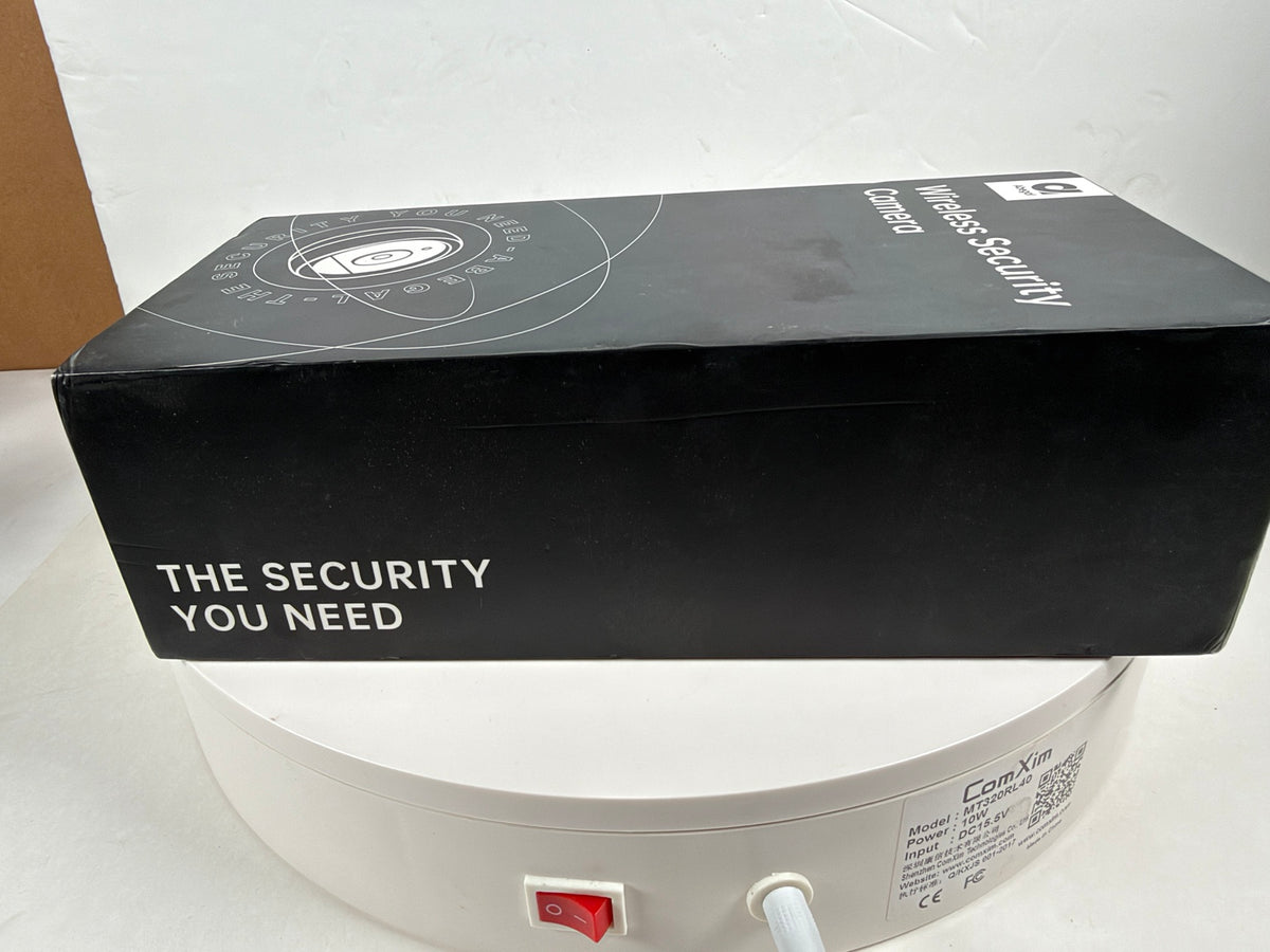 A black rectangular product box with white text reading 'THE SECURITY YOU NEED' and 'Wireless Security' sits on top of a white round base or stand with a red switch and product label visible.